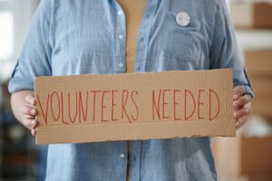 The photo is of the torso of a person, holding a "Volunteers Needed" sign.